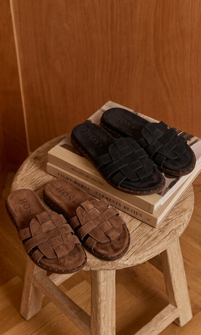Pair of brown sandals and a black sandal on a wooden stool with a wooden background.